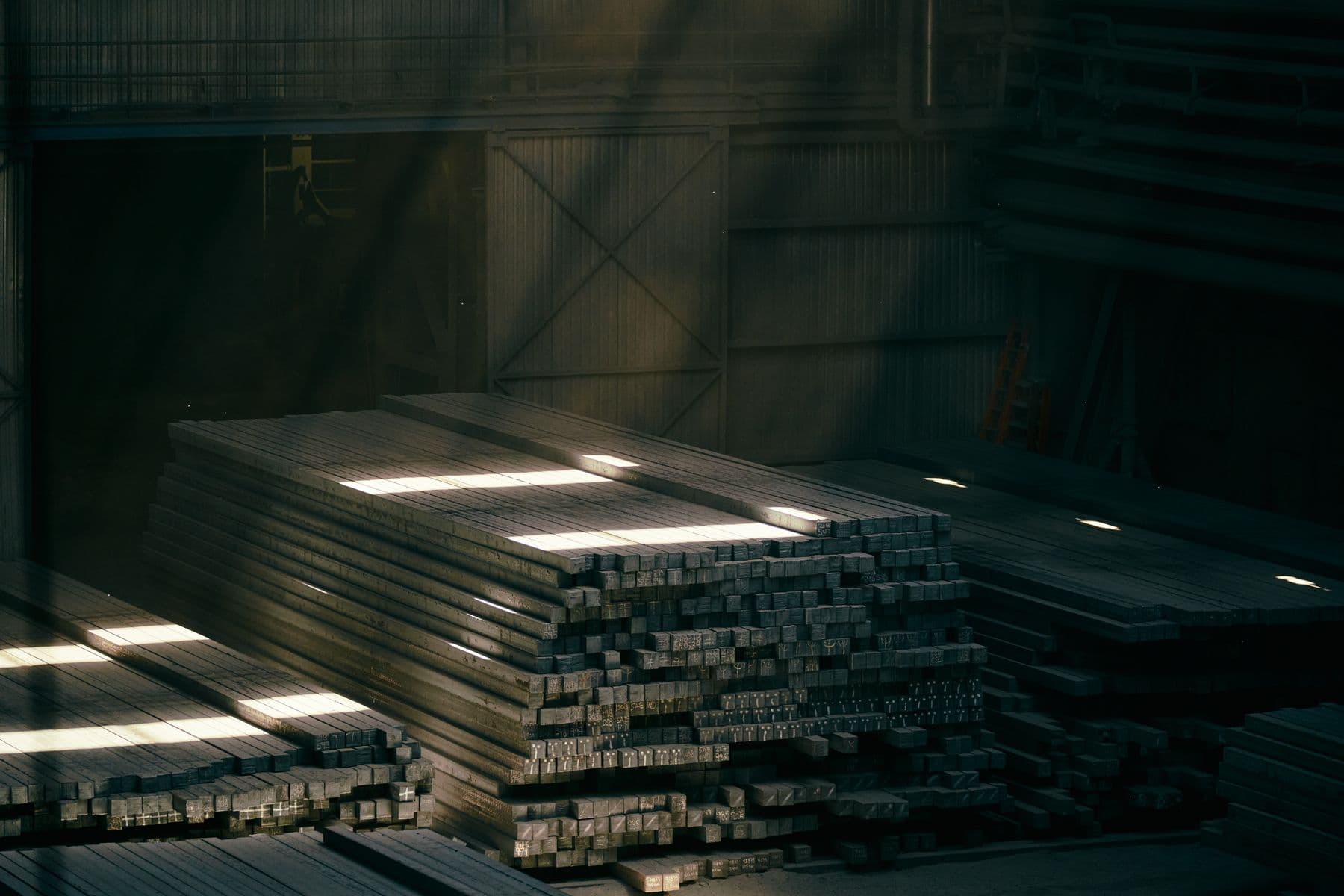 Piles de billettes et de plats d’acier dans un dépôt, éclairées par des rais de lumière.